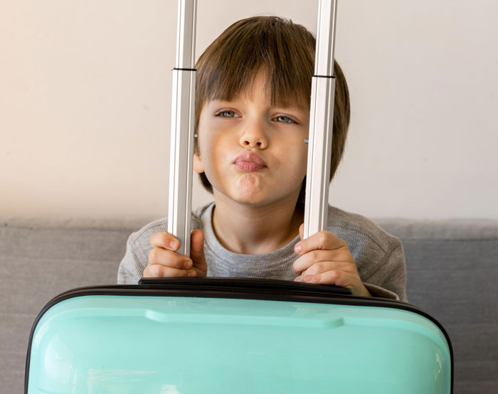 Young boy holding suitcase handle, preparing for trip, with mom planning Europe travel and ex requesting AirTag tracking.