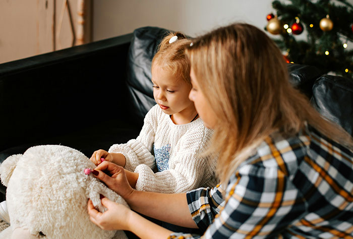A mom helps her child dress a stuffed animal, with a Christmas tree in the background.