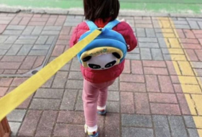 Child with panda backpack on a leash, wearing a red jacket and pink pants, walking on a paved sidewalk.