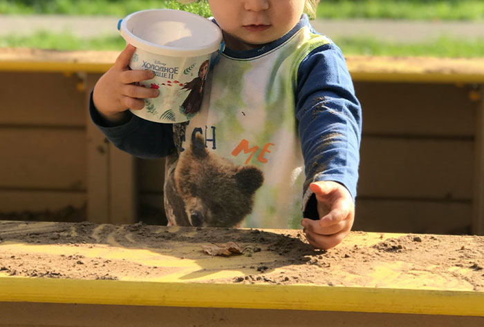 Toddler playing in sandbox, wearing a bear-themed outfit, with a container, illustrating stories of moms getting shamed.