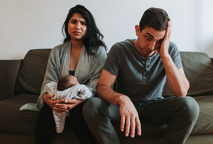 A mom with a baby and a concerned dad sitting on a couch, illustrating parenting challenges and mom shaming.