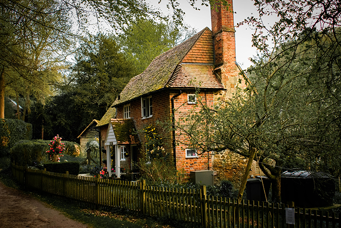 Cozy brick countryside house surrounded by greenery and a wooden fence on a peaceful tree-lined path.
