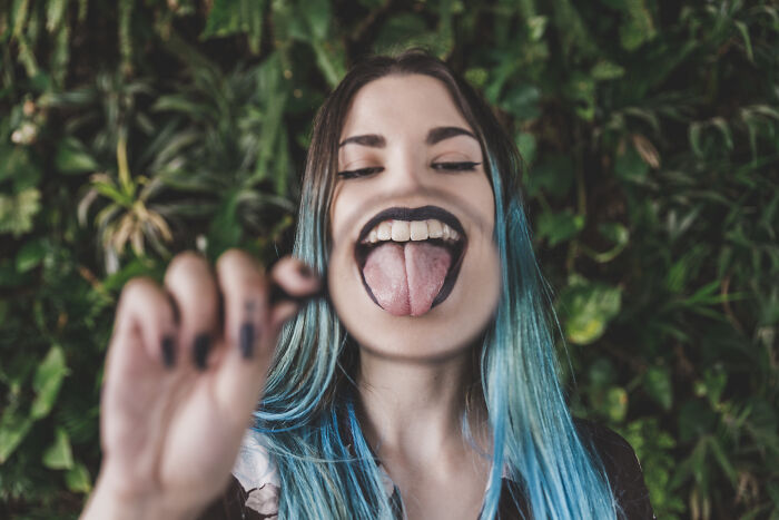 A person with blue hair making a silly face in front of a leafy background, embodying cringiest behavior.