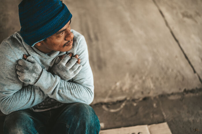 Man in blue beanie and gloves sits on concrete floor, embracing himself.