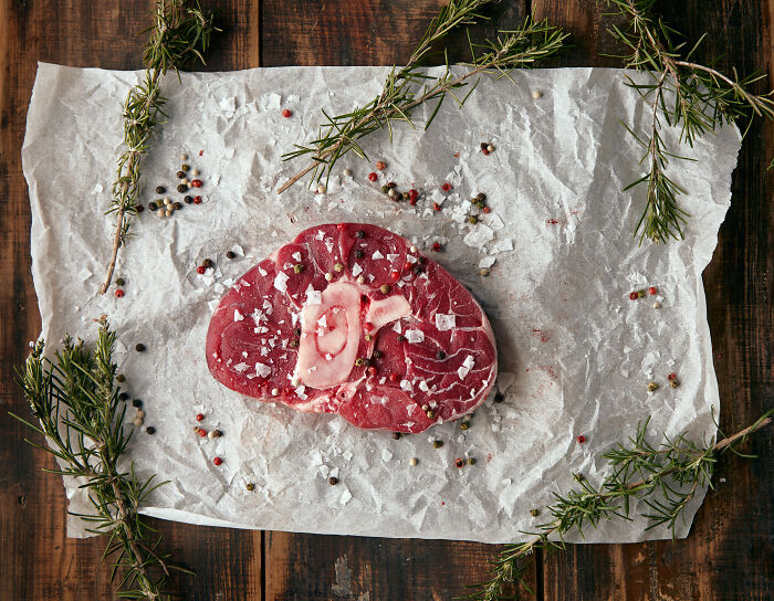 Raw steak on parchment paper, garnished with rosemary and pepper, showcasing a cringy food presentation.