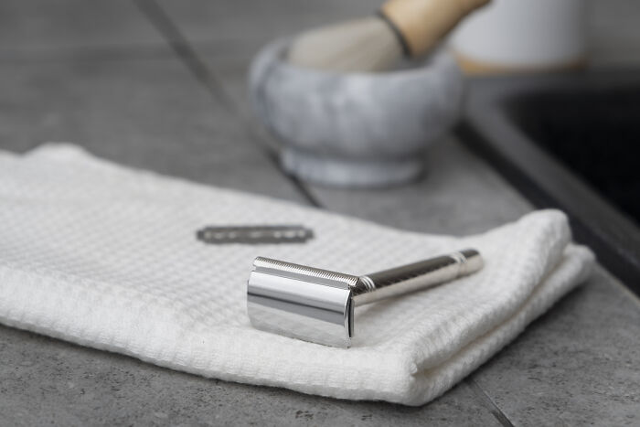 A shiny razor and a blade on a white towel, with a shaving brush in the background, exemplifying personal grooming tools.