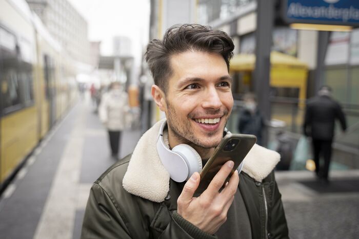Man smiling while talking into phone, wearing headphones, on a city street.