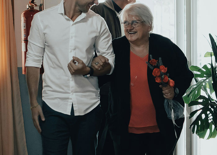 Elderly woman holding flowers, smiling and walking arm-in-arm with a younger man.