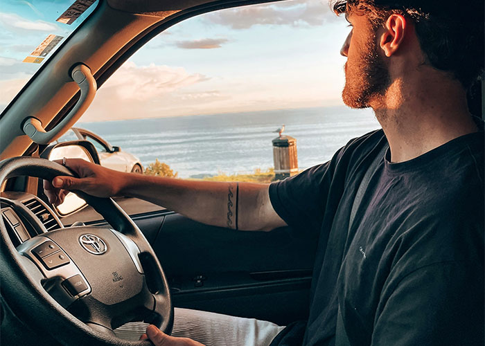 Man in a car looking at the ocean, gripping the steering wheel, with a tattoo on his arm, showcasing a serene moment.