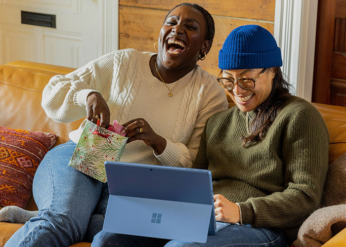 Two people laughing together on a couch, one holding a gift bag and the other using a laptop.