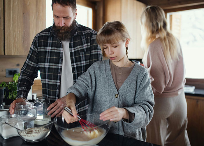 Family in kitchen, young girl whisking batter, father assisting, mother in background, highlighting family dynamics.