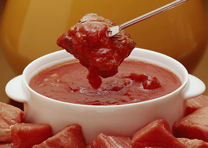 Piece of meat being dipped into a bowl of red sauce, surrounded by more raw meat.