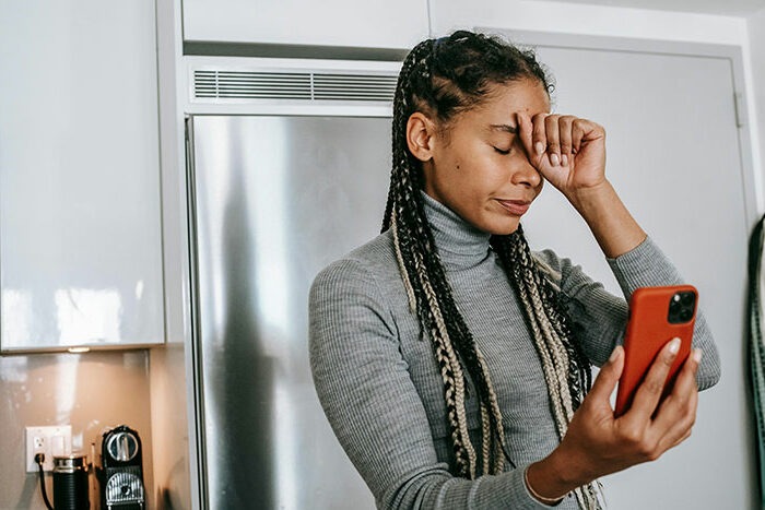 Woman feeling stressed in kitchen while looking at phone, highlighting family and support challenges.