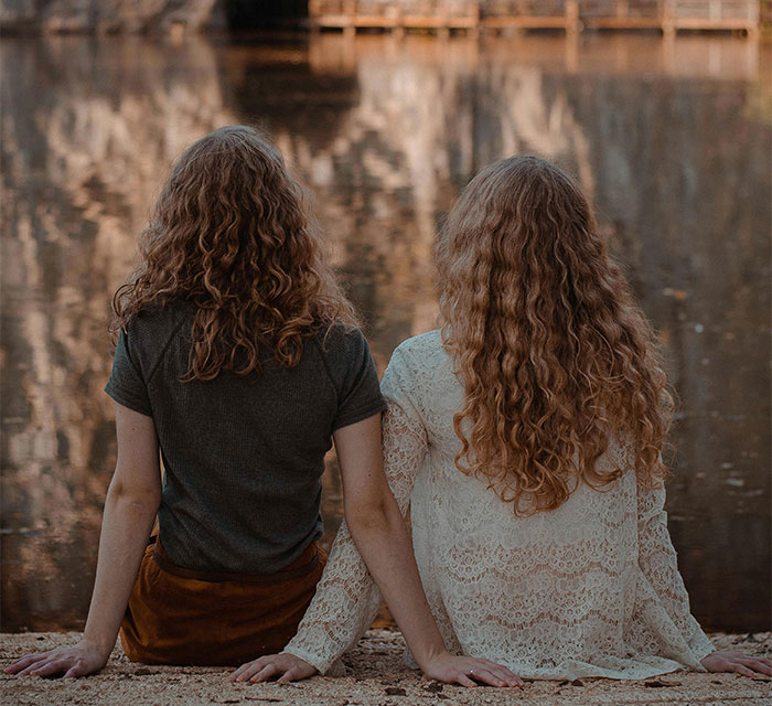 Two women with curly hair sit by a lake, facing away, symbolizing distance and family expectations.