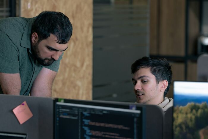 Two coworkers discussing code at a desk in an office setting.