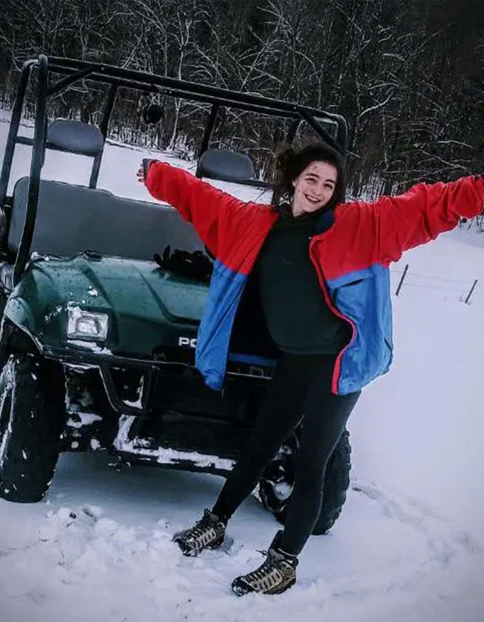 Former child star in snow, smiling next to a snow-covered vehicle, wearing a red and blue jacket.