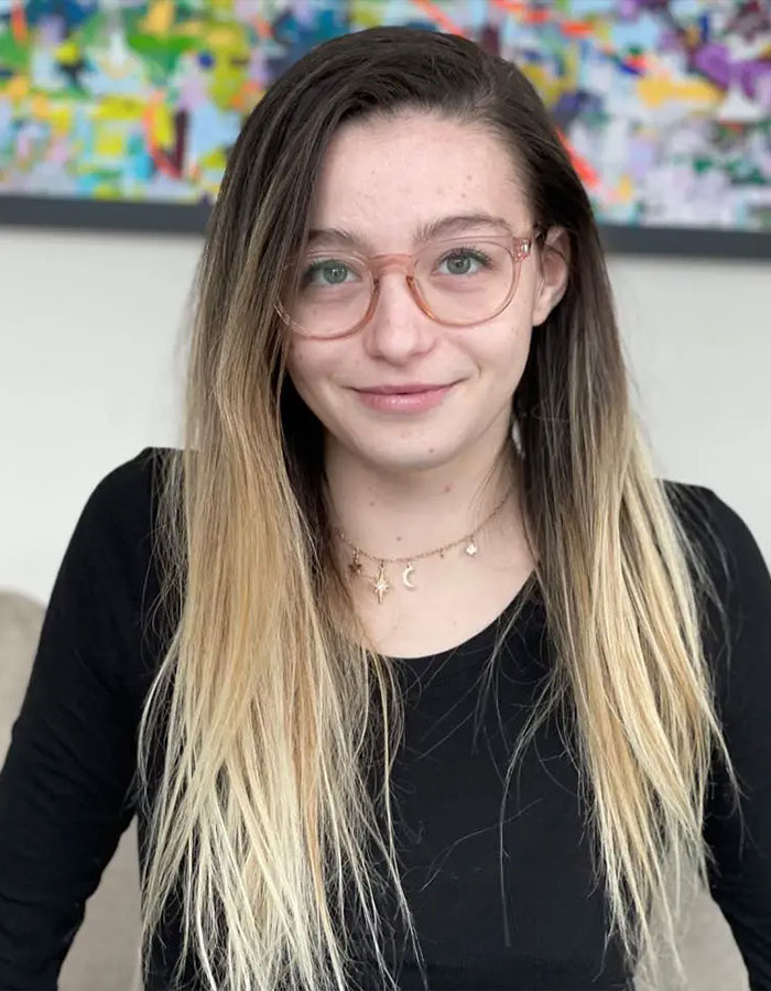 Young woman with long blond hair and glasses, wearing a black top, seated indoors with a colorful painting in background.