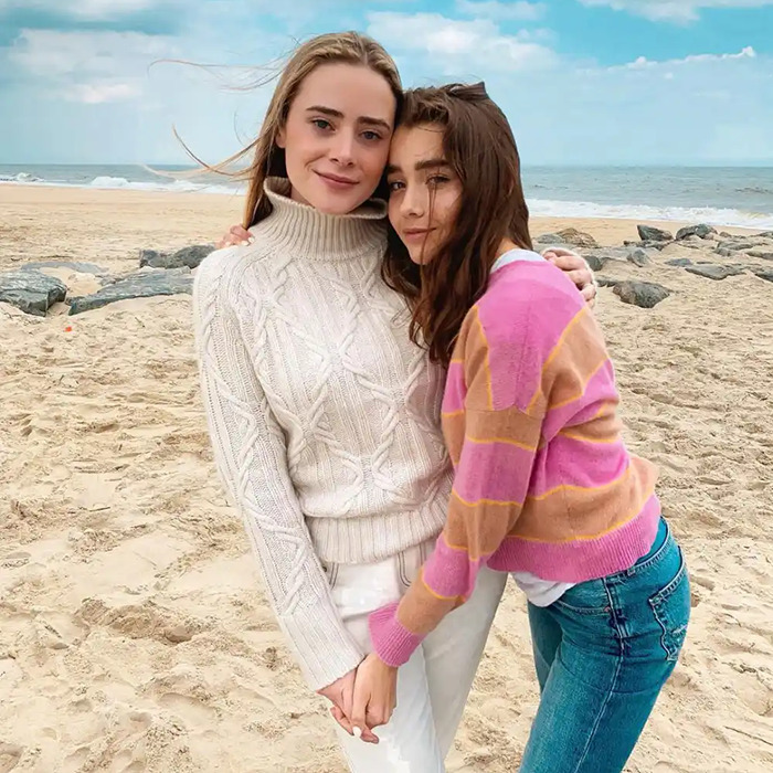 Two people on a beach, one in a cream sweater and the other in a pink striped sweater, embracing with the ocean behind.