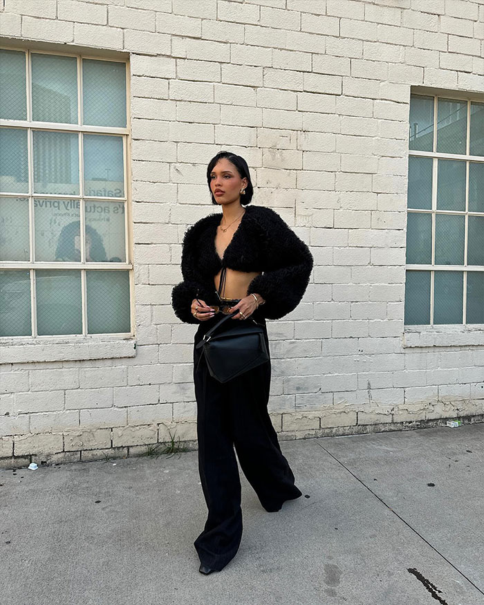 Nara Smith in stylish black outfit stands confidently against a brick wall, embodying calm and composure.