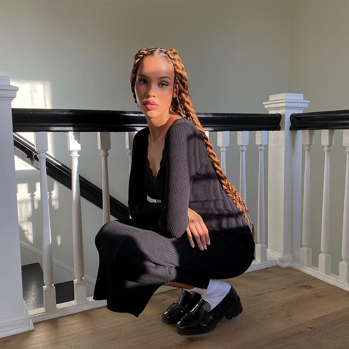 Nara Smith with long braids sitting on stairs, wearing a black dress, sunlight streaming in from the side.