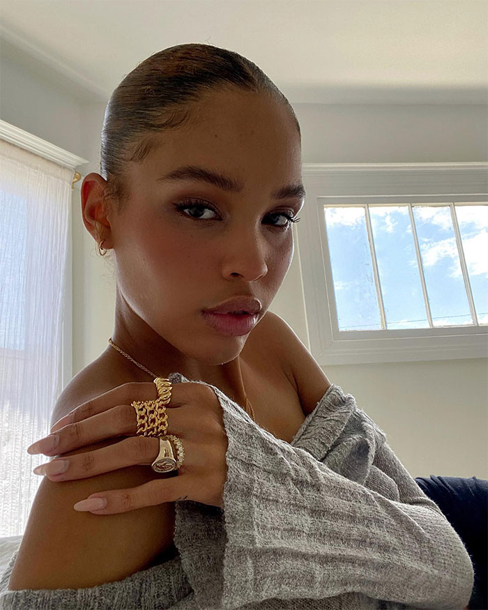 Nara Smith with poised expression, wearing a gray sweater and gold rings, sitting in a well-lit room.