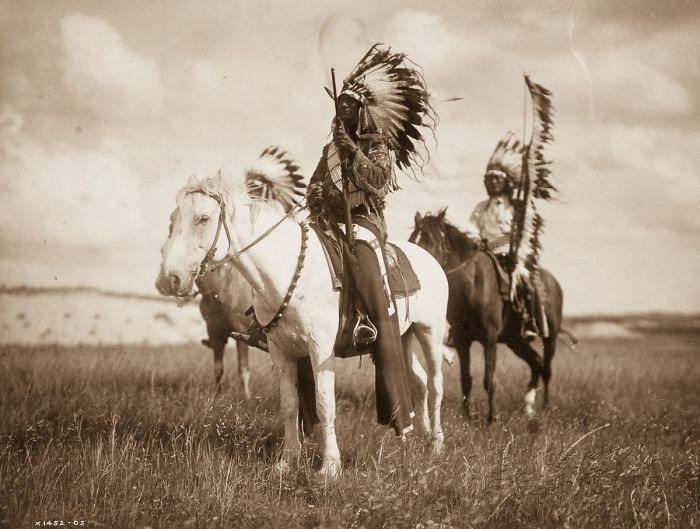 Native American men in traditional headdresses riding horses on a grassy plain, a historic photo revealing real lives.
