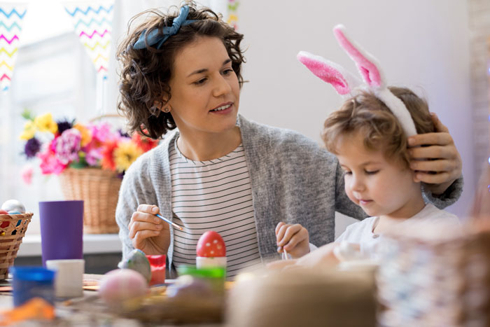 Woman and child with bunny ears painting Easter eggs in a bright, festive setting.