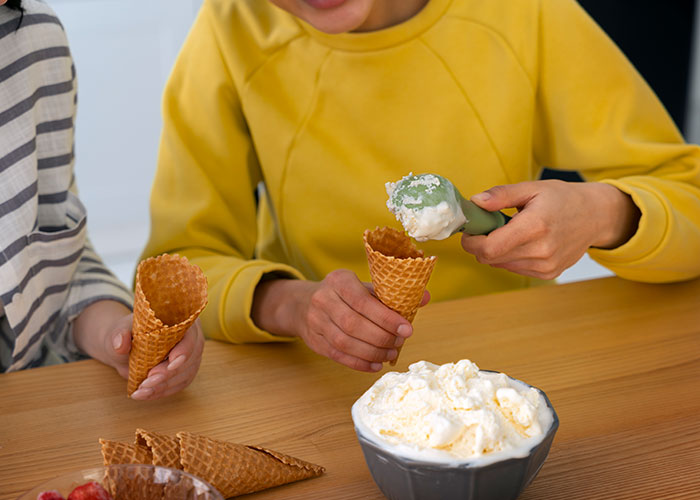 Child in yellow shirt scooping ice cream into a cone at table, with extra cones nearby.
