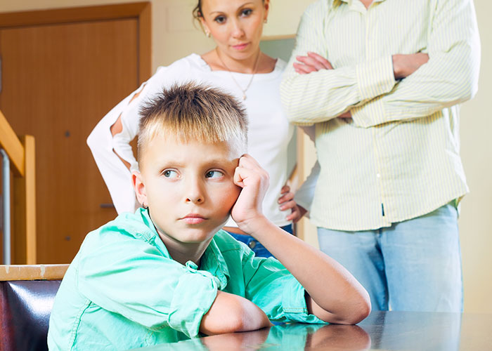A disappointed boy sits at a table, surrounded by two adults, looking upset.