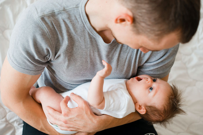 Father holding newborn baby, smiling in a cozy bedroom setting.