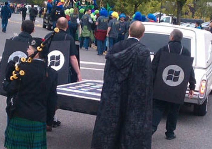 People in a parade, dressed in black with Windows logos, highlight tech flop in a humorous setting.