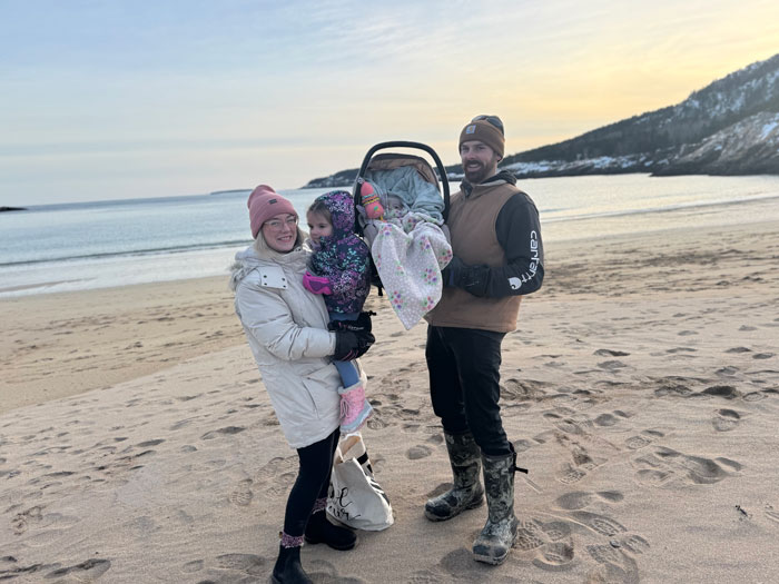 Influencer's family on a beach, holding a baby carrier in memory of their 10-month-old daughter.