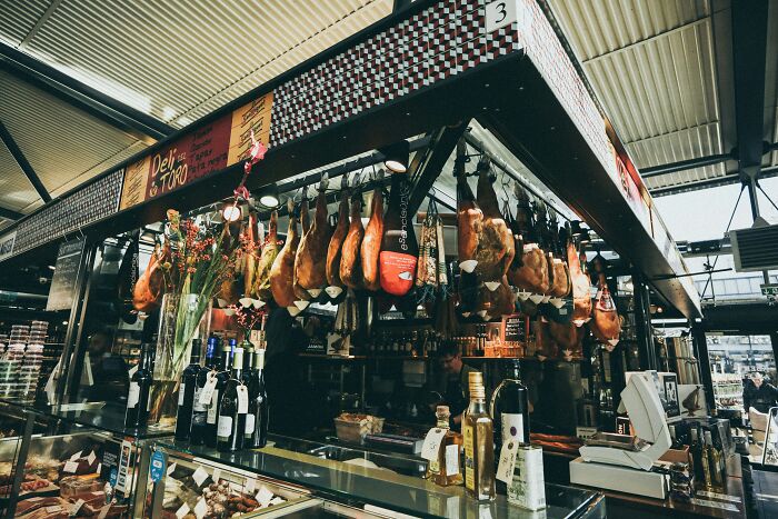 Deli market stall with hanging meats, wine bottles, and olive oil display, showcasing lively market ambiance.