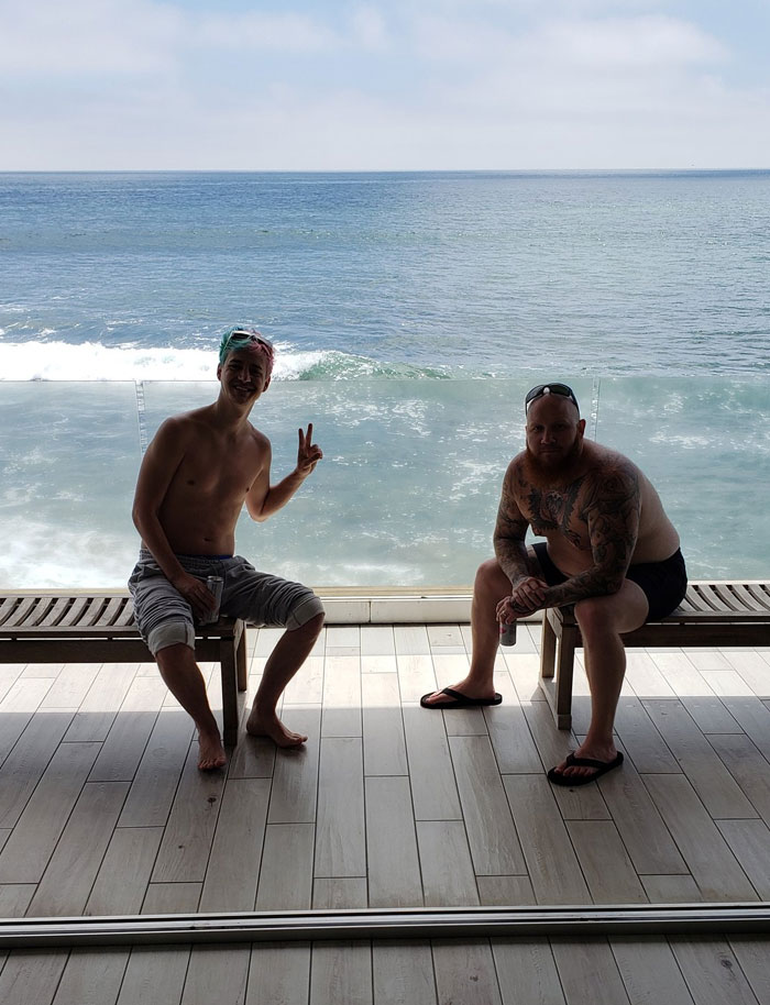 Two people including Ninja sitting by the ocean, Ninja giving a peace sign, embodying relaxation and lifestyle of a streaming legend.