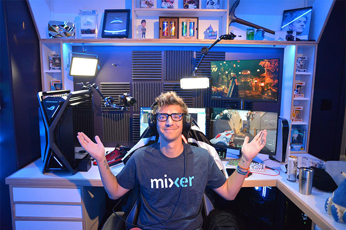 Streamer Ninja in a gaming room, wearing a "Mixer" shirt and glasses, surrounded by monitors and lighting equipment.