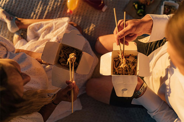 Two people enjoying takeout with chopsticks, possibly embracing a "No-Buy" lifestyle to resist consumerism.