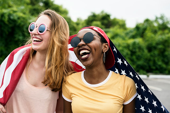 Two friends smiling outdoors with an American flag, enjoying US culture and travel experiences together.