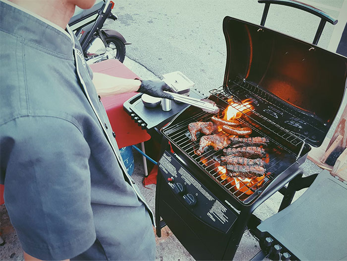 Person grilling assorted meats on a gas barbecue, showcasing popular US culture outdoor cooking and dining tradition.