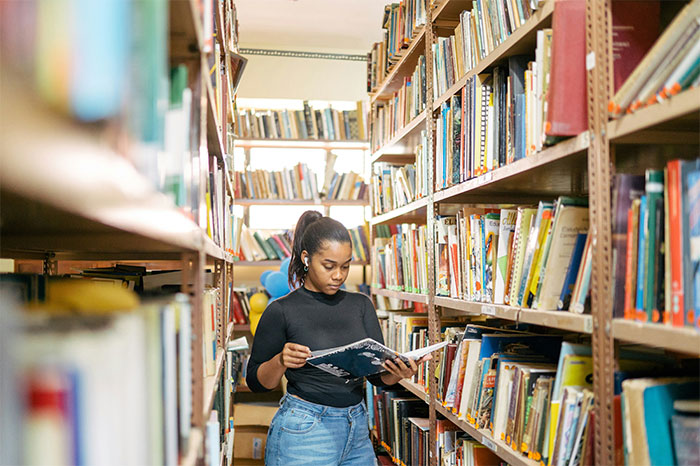 Young woman reading a book in a library aisle, exploring nuances about US culture during her travels.