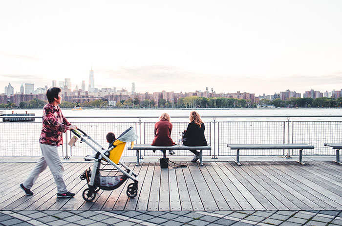 Person pushing stroller on a wooden boardwalk with two people sitting by the river, reflecting US culture nuances.
