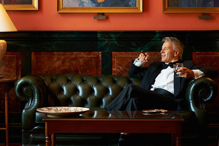 Man in tuxedo relaxing on a leather couch, enjoying a cigar and drink, reflecting US culture nuances for travelers.