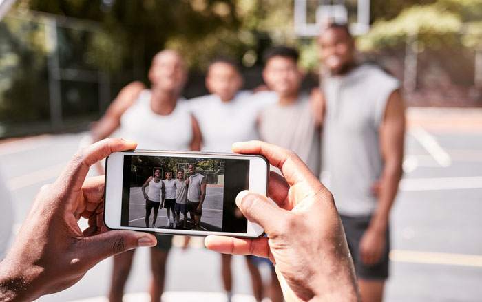 Group photo of friends on a basketball court, focus on phone capturing the image, causing surprise among onlookers. Group photo of friends on a basketball court, focus on phone capturing the image, causing surprise among onlookers.