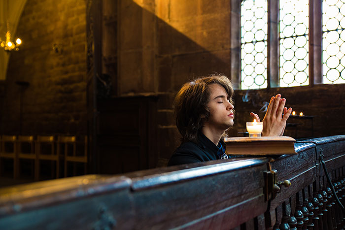 A person in medieval attire praying near a candle in a dimly lit, rustic church setting.