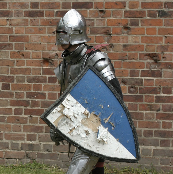 Medieval knight in armor with a blue shield, standing against an old brick wall.