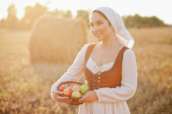 A woman in medieval attire holds a basket of apples in a sunny field, symbolizing love potions from times past.