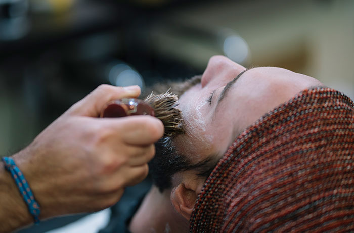Man getting a shave at barbershop, lying down with barber using brush on face, medieval grooming style.
