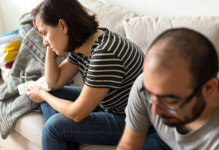 Wife and husband sitting on a couch, looking upset after a discussion involving his female best friend and their child&rsquo;s birth.
