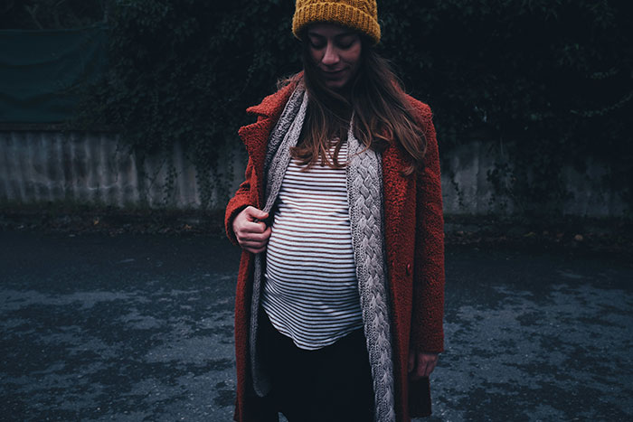 Pregnant woman in a striped shirt and orange coat, outdoors on a dimly lit road.