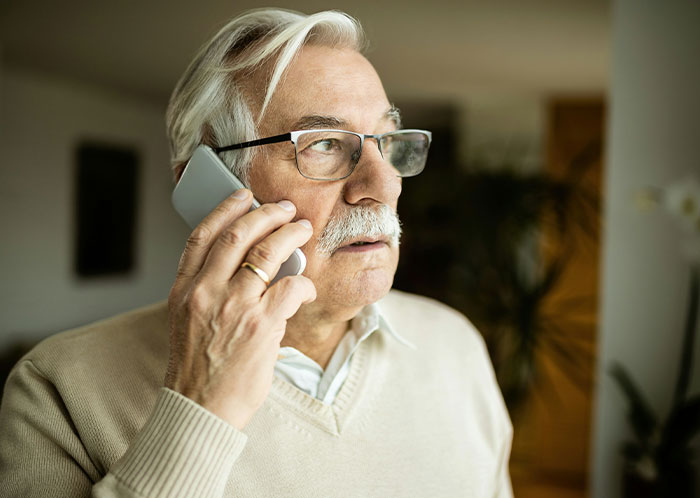 Elderly man with glasses on phone, appearing upset, in a beige sweater.