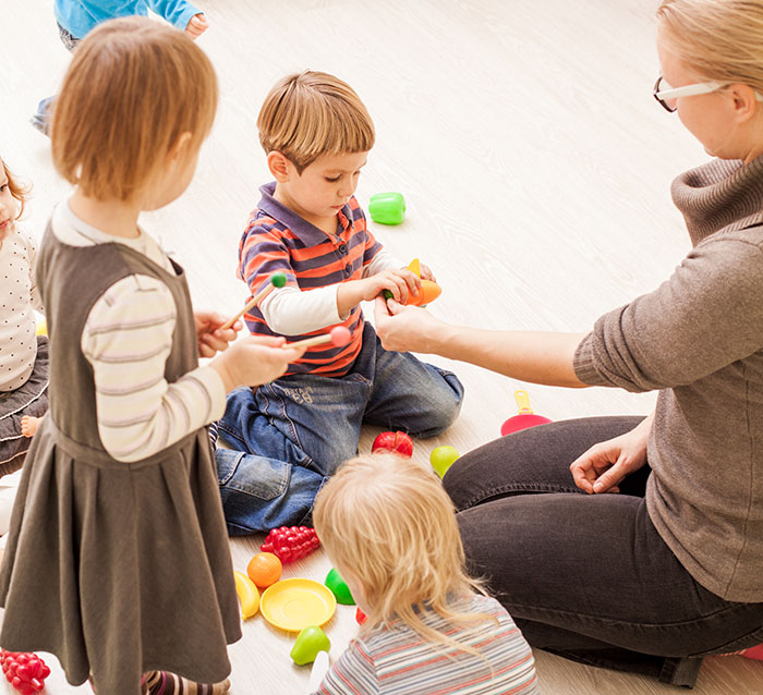 Children playing with colorful toys in a classroom setting, engaging with a teacher.